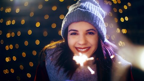 Cute girl with sparkling stick smiling into the camera. Portrait sliding shot of young woman in focus wearing winter clothes and surrounded by decorative lights.