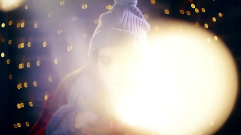 Girl opens the Christmas present with big smile. Portrait sliding shot of young woman in focus wearing winter clothes and surrounded by decorative lights.