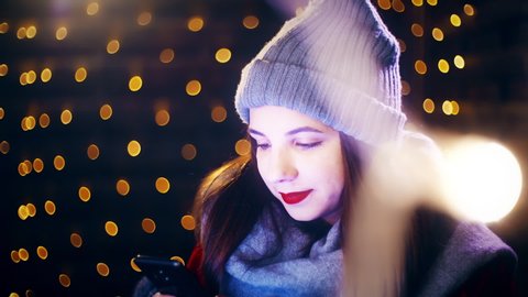 Girl on the smartphone while surrounded by Christmas lights. Portrait sliding shot of young woman in focus wearing winter clothes and surrounded by decorative lights.