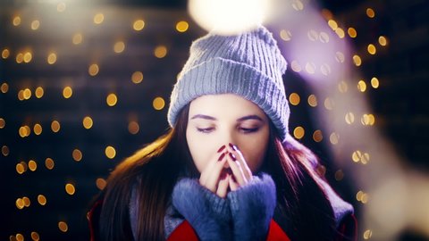 Girl with cold fingers outside surrounded by Christmas lighths. Portrait sliding shot of young woman in focus wearing winter clothes and surrounded by decorative lights.