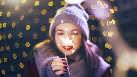 Person hold sparkler in Christmas spirit. Portrait sliding shot of young woman in focus wearing winter clothes and surrounded by decorative lights.