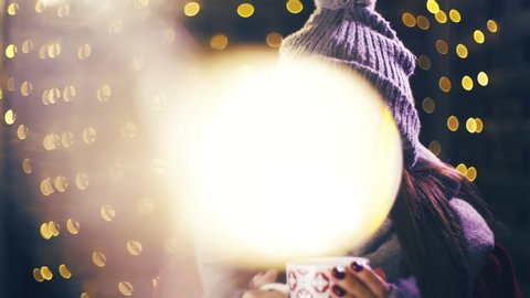 Young woman with a cup of warm drink in hands for Christmas. Portrait sliding shot of young woman in focus wearing winter clothes and surrounded by decorative lights.