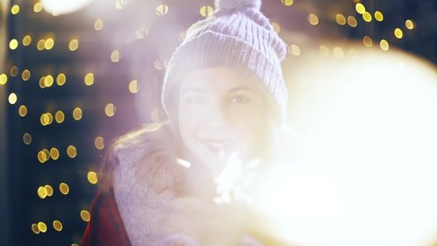 Cute girl portrait with sparkler looking into camera. Portrait sliding shot of young woman in focus wearing winter clothes and surrounded by decorative lights.