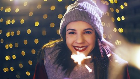 Girl with a sparkler enjoying time for Christmas. Portrait sliding shot of young woman in focus wearing winter clothes and surrounded by decorative lights.