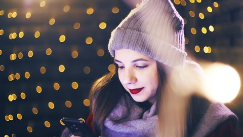 Lonesome young woman on the smartphone for Christmas. Portrait sliding shot of young woman in focus wearing winter clothes and surrounded by decorative lights.