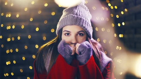 Girl with a cute look freezing outside for Christmas. Portrait sliding shot of young woman in focus wearing winter clothes and surrounded by decorative lights.