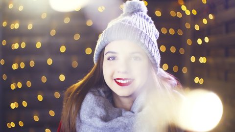 Portrait of attractive girl in warm clothes surrounded by Christmas lights. Portrait sliding shot of young woman in focus wearing winter clothes and surrounded by decorative lights.