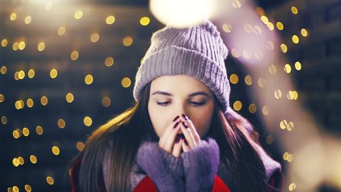 Girl warming her beautiful fingers while freezing outside. Portrait sliding shot of young woman in focus wearing winter clothes and surrounded by decorative lights.