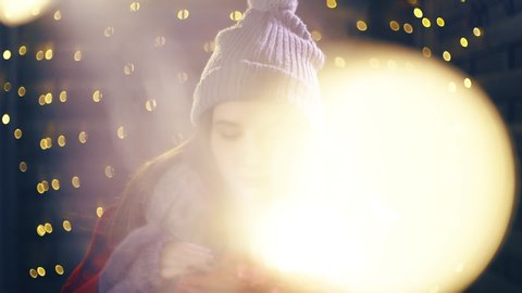 Christmas gift shines over the face of cute girl smile. Portrait sliding shot of young woman in focus wearing winter clothes and surrounded by decorative lights.