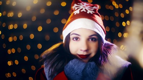 Christmas cute girl looking at the camera. Portrait sliding shot of young woman in focus wearing winter clothes and surrounded by decorative lights.