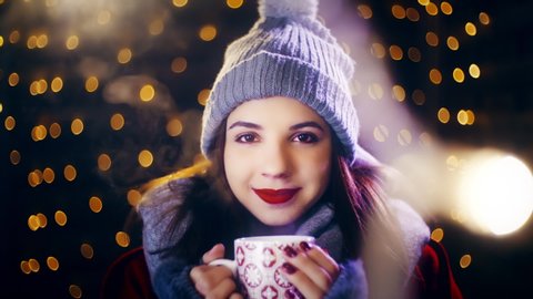 Girl enjoying cup of warm drink for Christmas. Portrait sliding shot of young woman in focus wearing winter clothes and surrounded by decorative lights.