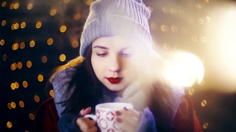 Christmas cute girl holding a cup of drink and smile. Portrait sliding shot of young woman in focus wearing winter clothes and surrounded by decorative lights.