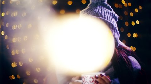 Girl blows the hot drink in hands surrounded by Christmas lights. Portrait sliding shot of young woman in focus wearing winter clothes and surrounded by decorative lights.
