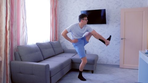 Funny nerd man in glasses and shorts is doing stretching exercise for leg, trying to lift his leg up, and falling down the floor at home. Sport humor concept.