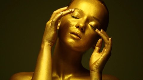 Fashion art Golden skin Woman face portrait closeup. Model girl with holiday golden Glamour shiny professional makeup. Gold jewellery, jewelry, accessories. Beauty gold metallic body, painted Skin 4K
