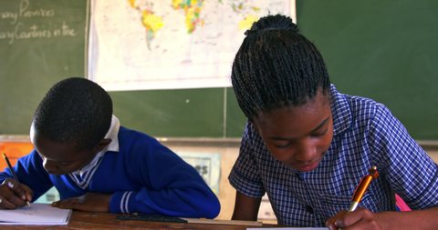 Front view close up of a young African schoolgirl and schoolboy sitting at desks smiling, writing in her note book and listening attentively during a lesson in a township elementary school classroomの動画素材
