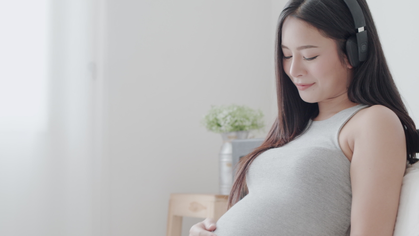 Pregnant Asian mother sitting   open music headset put hand her tummy with soft touch to make her child feeling relaxed on bedroom in the morning time | Shutterstock HD Video #1035436610