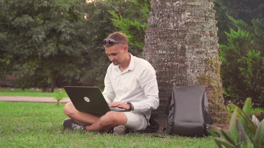A young man sits in a Park on the grass near a palm tree and prints a text on a laptop. A man works on a laptop outside the house.