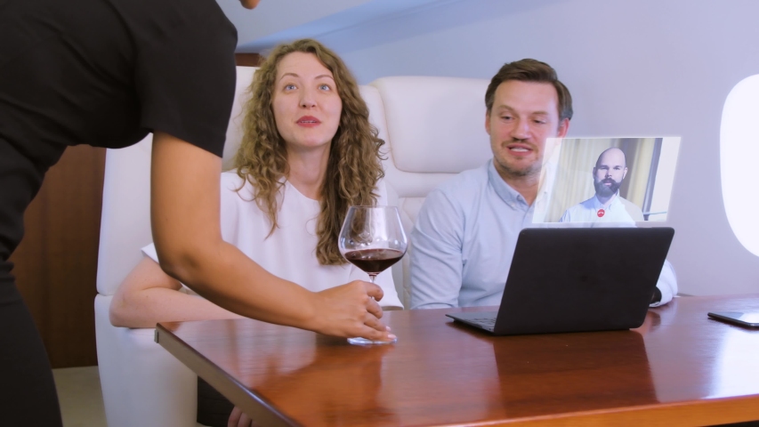 Making video conference call with colleague on laptop HUD hologram on board of private jet. Flight attendant offer glass of wine for caucasian businessman and businesswoman inside business airplane - Powered by Shutterstock - Get 15% off with code: PIKWIZARD15