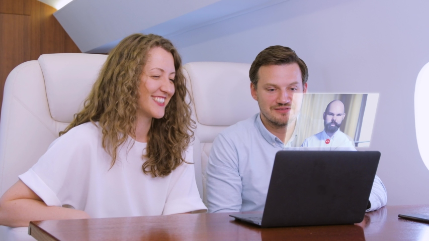 Making video conference call with colleague on laptop HUD hologram on board of private jet. Caucasian businessman and businesswoman travel inside of business airplane cabin. - Powered by Shutterstock - Get 15% off with code: PIKWIZARD15