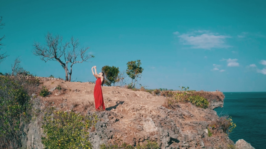 Aerial drone view of beautiful woman in red dress posing on the sea shore cliff during sunny summer day
