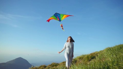 LGBT love and freedom. View of attractive girl activist walking on green mountain top holding colorful flying snake against blue sky background. Amazing scenic nature.