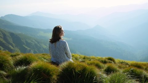 Enchanting girl in dress sitting on mountain top enjoying summer nature scenery. Happy relaxed girl with eyes closed dreaming thinking positive staying in highland. Green landscape, natural glory.