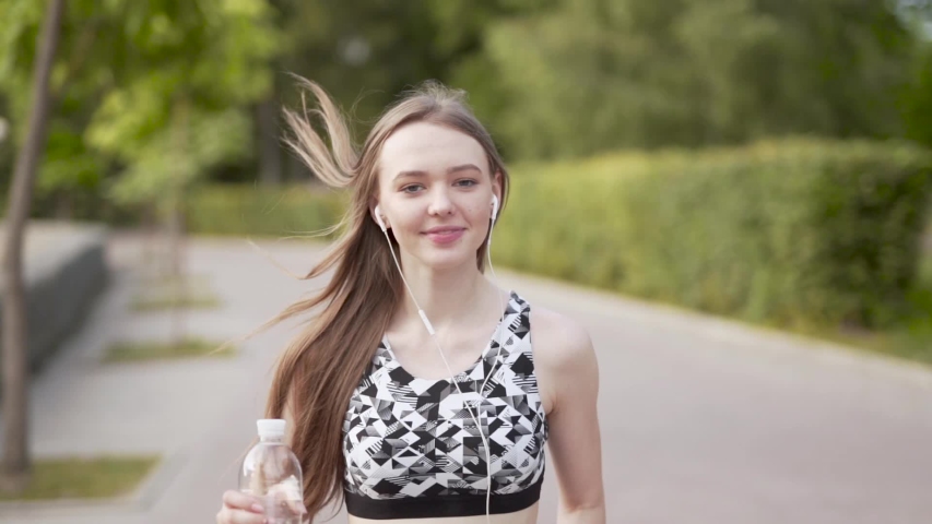 Young beautiful slender womn is running in the park. She has an earphones and running in front of camera.