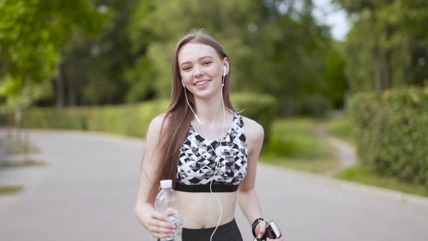 Young beautiful slender womn is running in the park. She has an earphones and running in front of camera.