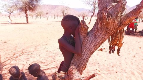 NAMIBIA - CIRCA 2018 - A young African Himba tribal boy leans against a tree in a small village in Namibia.
