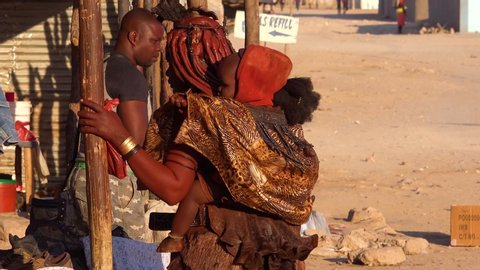 OPUWO, NAMIBIA - CIRCA 2018 - A Himba tribal woman with baby on back and amazing hairstyle of mud and braids and dreadlocks.