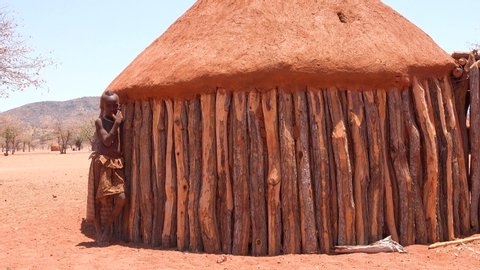 NAMIBIA - CIRCA 2018 - A young African Himba tribal boy leans against his mud and wood hut in a small village in Namibia.