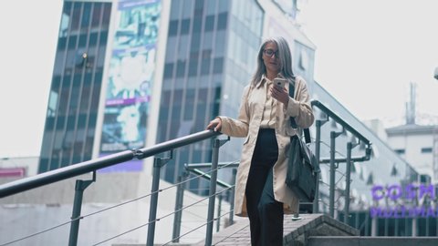 The Middle-Aged Asian Woman In A Fashionable Coat , A Shirt And Black Trousers Is Walking Down The Stairs. She Looks At The Screen Of Her Mobile Telephone And Types. Sometimes She Stops And Looks Away