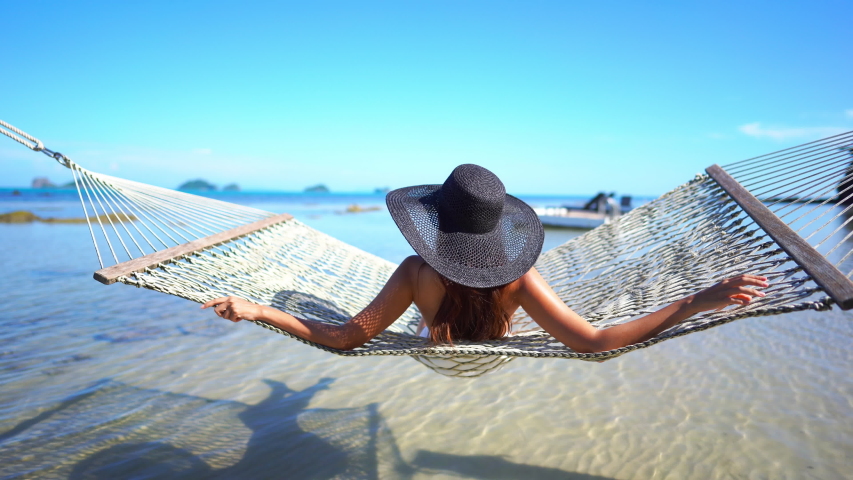 Young woman wearing a floppy black straw hat sitting in the sun and swinging in a hammock while looking out at the ocean.