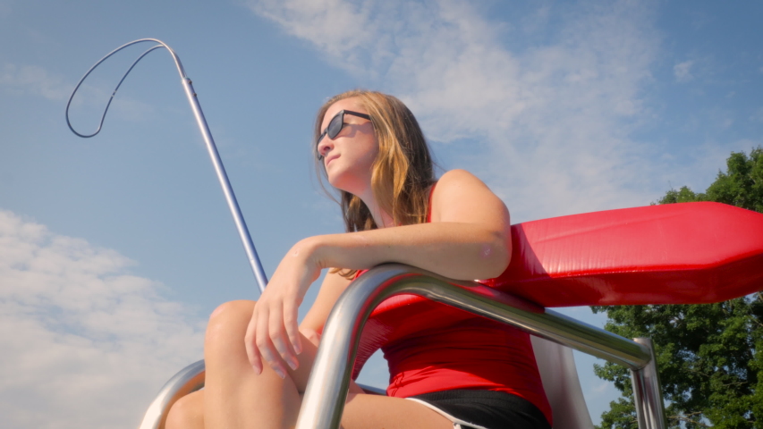 Low angle of a female lifeguard on a stand watching over swimmers at a public pool. The teen blows her whistle while working a summer job lifeguarding. Action shot represents employment and safety. Royalty-Free Stock Footage #1035486194