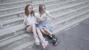 Two young sporty girls sitting on steps dancing to music with roller skates