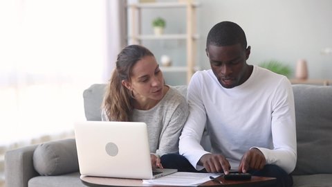 African husband biracial wife sit on couch at home planning family budget check online banking summary, calculate manage incomes and expenses use calculator and computer, check discuss loan agreement