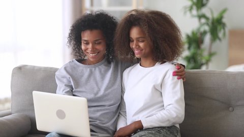 Mixed race mother sit with adolescent pre-teen grown-up daughter on sofa use notebook apps watching film online, internet users. Older younger sisters spend free time having fun with electronic device