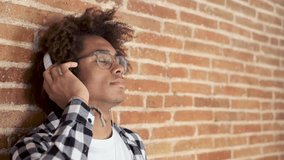 Video of relaxed afro-american young man listening to music with headphones leaning on stone wall at home.