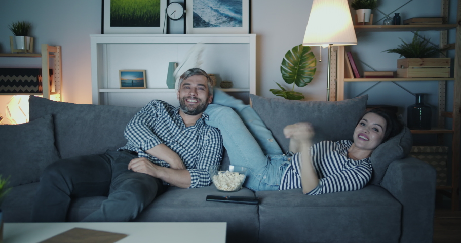Joyful man and woman are watcing TV throwing popcorn and laughing having fun together in house at night. Happy people, relationship and home concept. | Shutterstock HD Video #1035512450