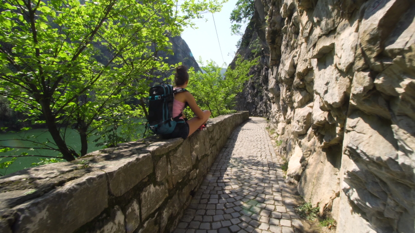 girl with backpack on cobbled pavement trail, enjoying the view of a river lake
