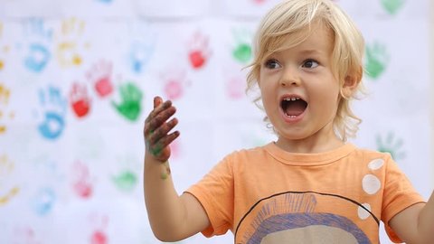 Amazing cute happy child surprising, saying wow and rubbing color paint on his little hands on color handprints background 50fps"