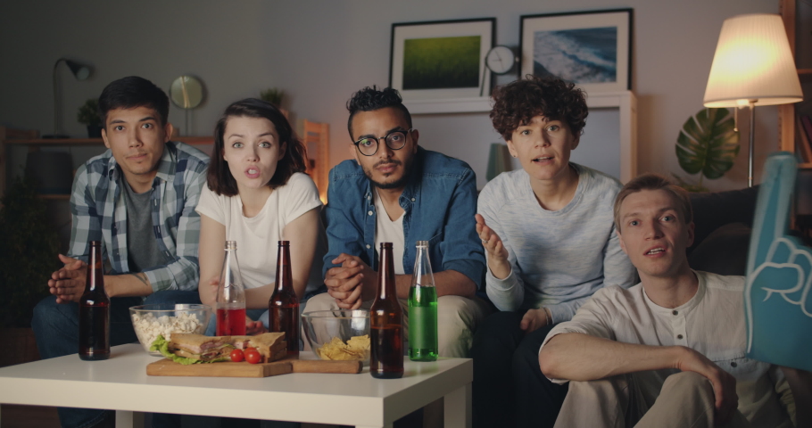 Portrait of happy young people watching sports game on TV at night cheering looking at camera sitting on couch together eating snacks and drinking