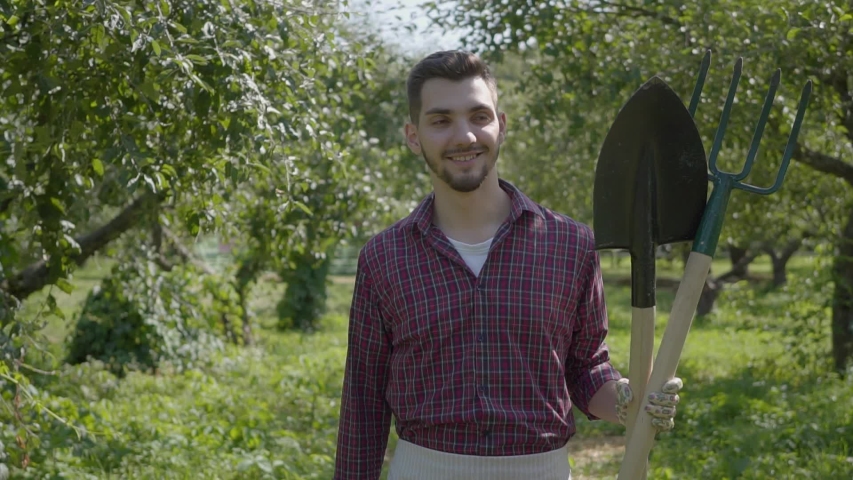 Young smiling bearded farmer walking through the garden with a shovel and pitchfork in hands. Concept of rural life, fruit-growing, gardening. Slow motion