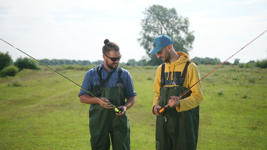 two men fisherman, with black hair and beard, green overalls, holding a fishing rod and a float, communicate with each other, smile, on the street, by the river on the grass, Sunny weather, close-up