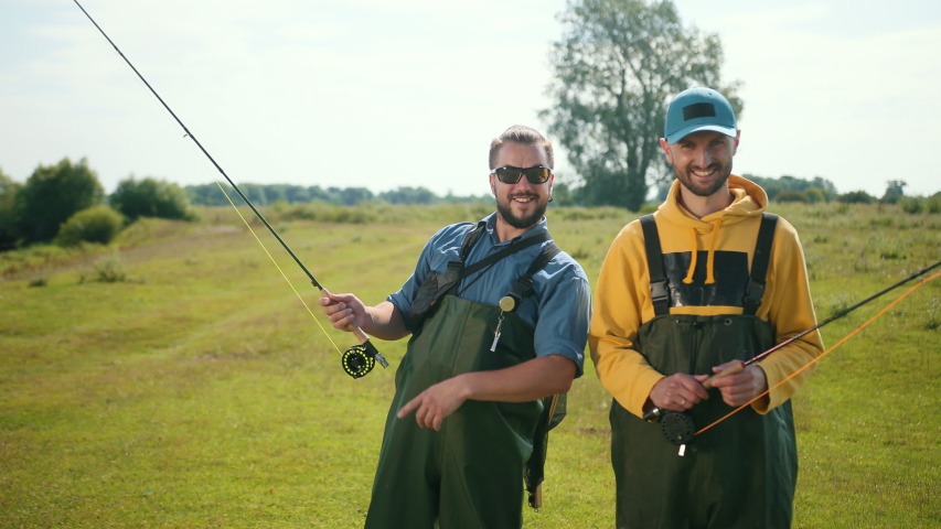 two men fisherman, with black hair and beard, green overalls, holding a fishing rod and a float, communicate with each other, smile, on the street, by the river on the grass, Sunny weather, close-up