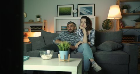 Young couple man and woman watching boring film on TV at home at night yawning relaxing on couch together with bored faces. People, house and television concept.