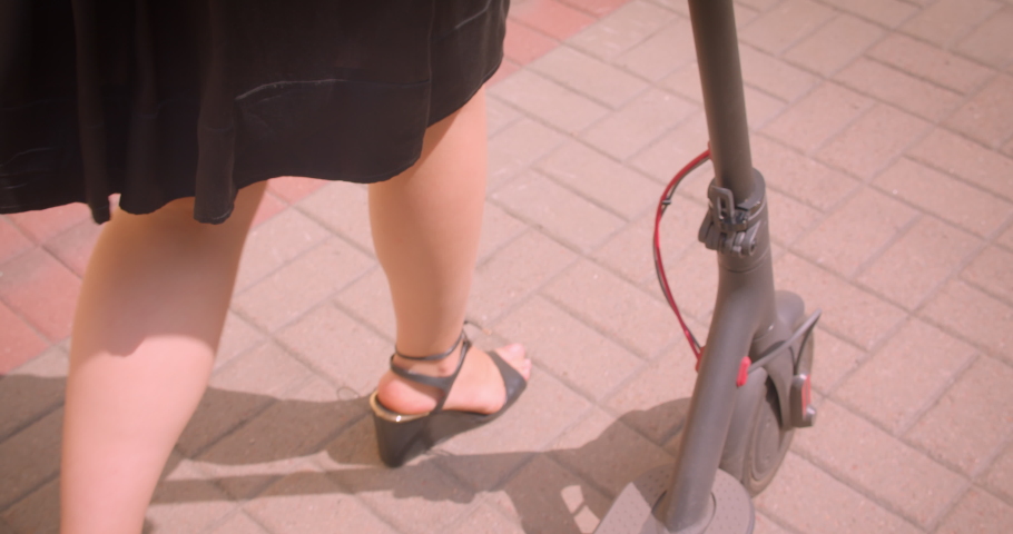 Closeup portrait of young beautiful female with long hair in glasses and dress walking with kick scooter happily outdoors