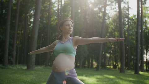 Pregnant woman doing yoga standing in warrior position in nature outdoors on sunny day. Young mother to be practice stretching yoga in green park with trees on background