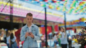stylish hipster in denim jacket holds white smartphone looks for friend smiles under colorful roof closeup slow motion